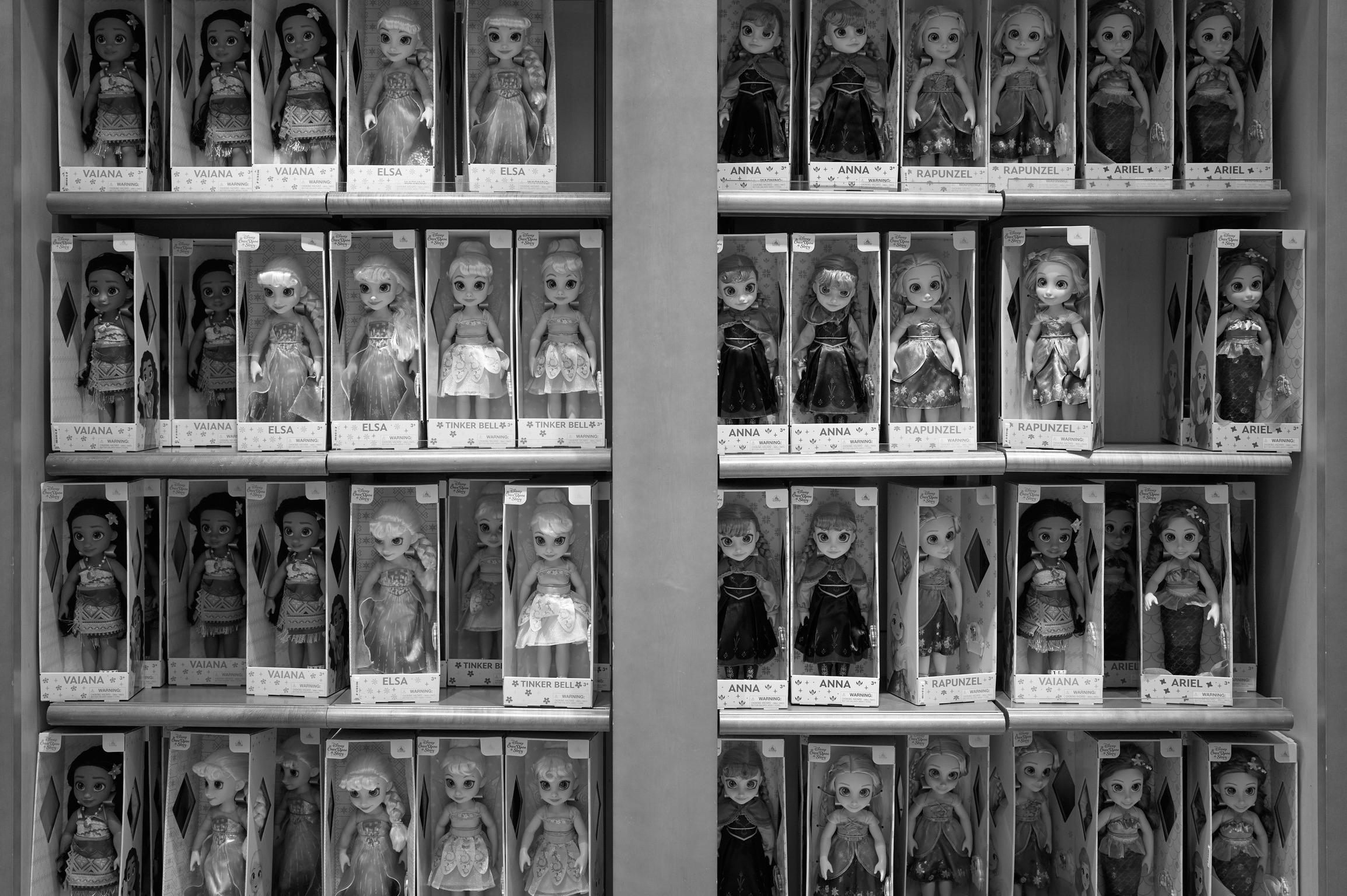 A display of various boxed dolls arranged on shelves, featuring characters like Elsa, Anna, Moana, Tinker Bell, Rapunzel, and Ariel. The image is in black and white.